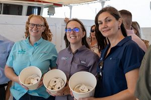 Three women stand on a boat holding containers filled with oysters.