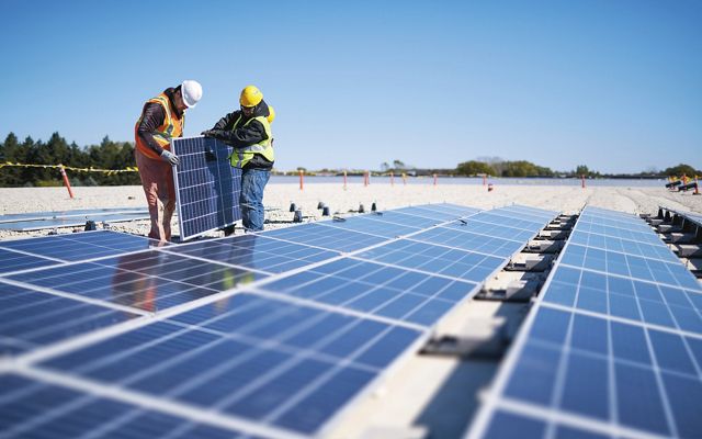 two workers carry a solar panel to be installed in a row of panels on dirt ground