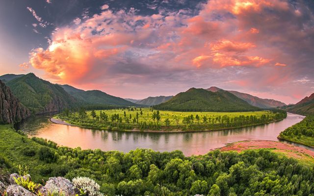 A wide river curves between lush green banks. Mountain peaks rise in the background under clouds that glow golden in the setting sun.