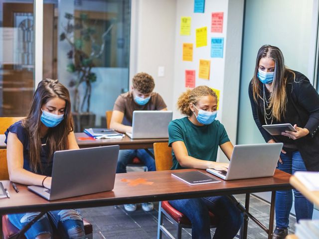 A teacher assists 3 socially-distancing and masked students with assignments as they work from their laptops in a classroom.