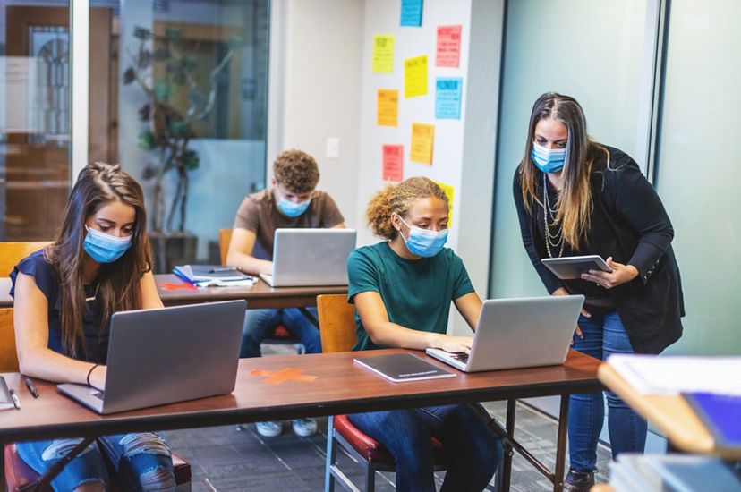 A teacher assists 3 socially-distancing and masked students with assignments as they work from their laptops in a classroom.