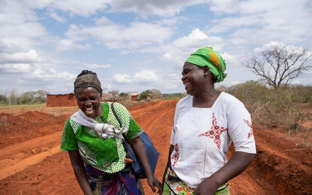 Two women laughing in Kenya while standing on a red clay path. 