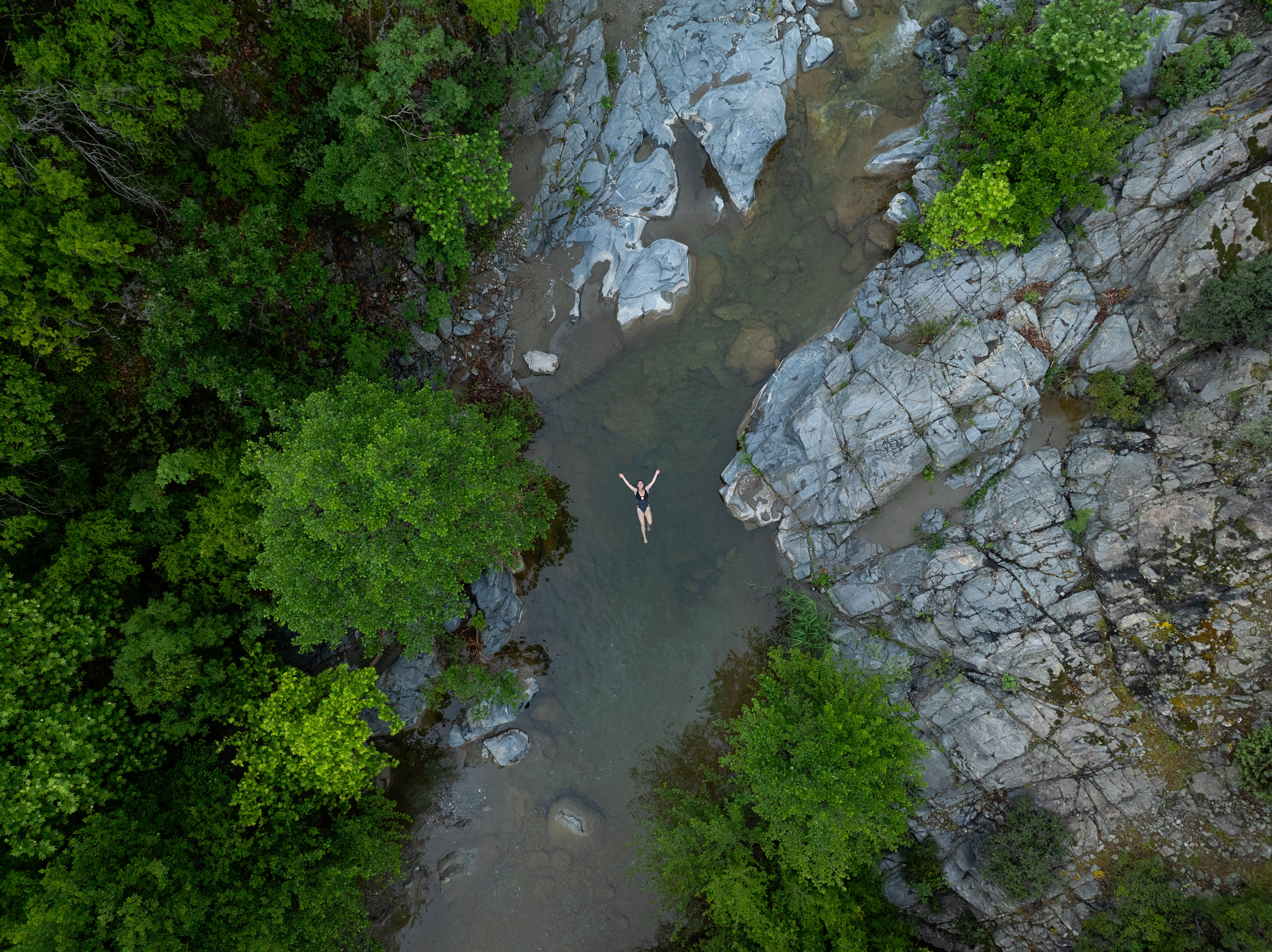 A view from above shows a woman swimming in a river with a rocky shoreline and forest beyond.