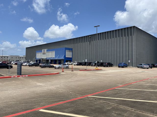 A metallic and mostly windowless rectangular building is surrounded by a large asphalt parking lot on a sunny day in Houston. The building is a high school.