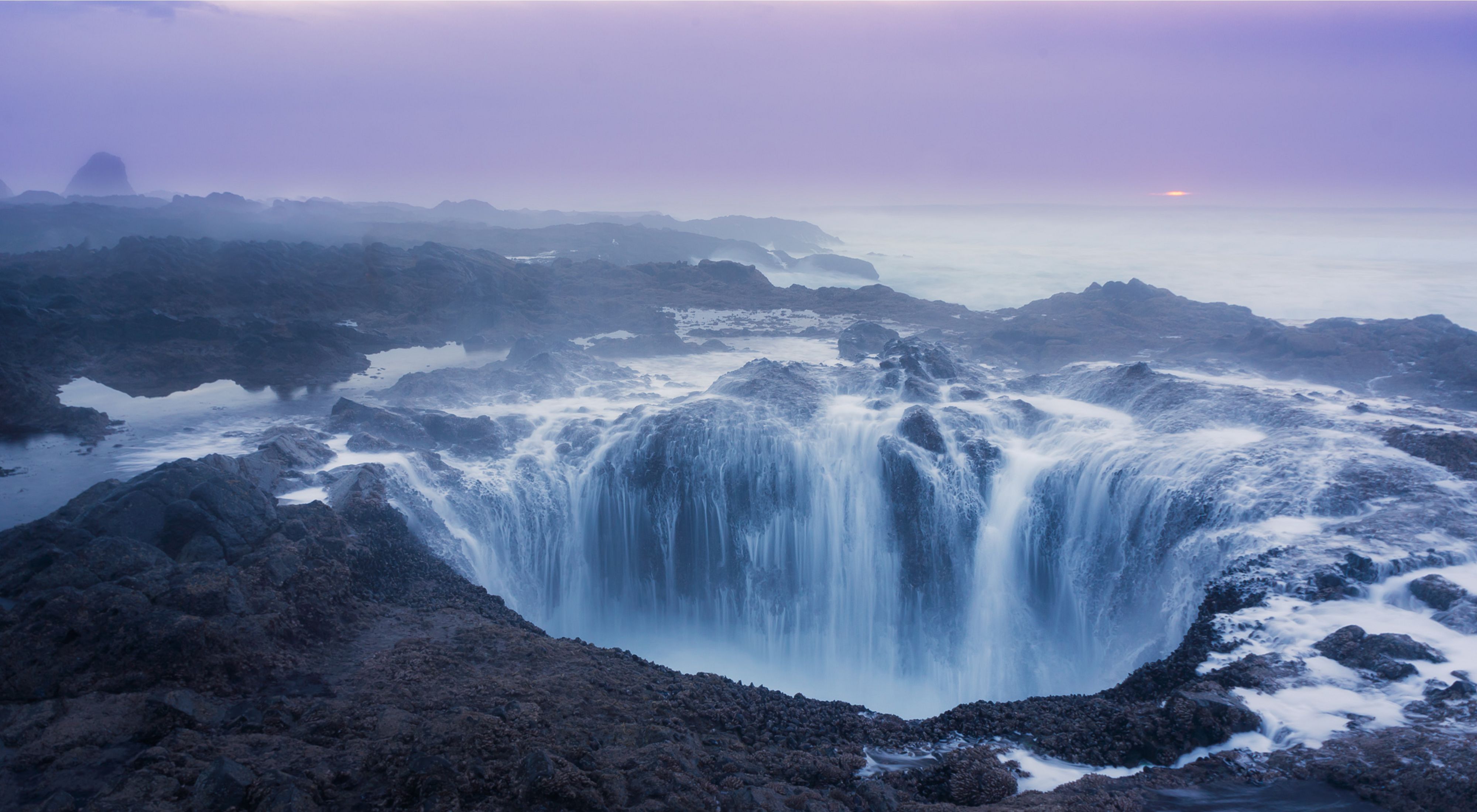 Tidal waters rushing into a large hole in black coastal rocks surrounded by blue sea and purple skies.