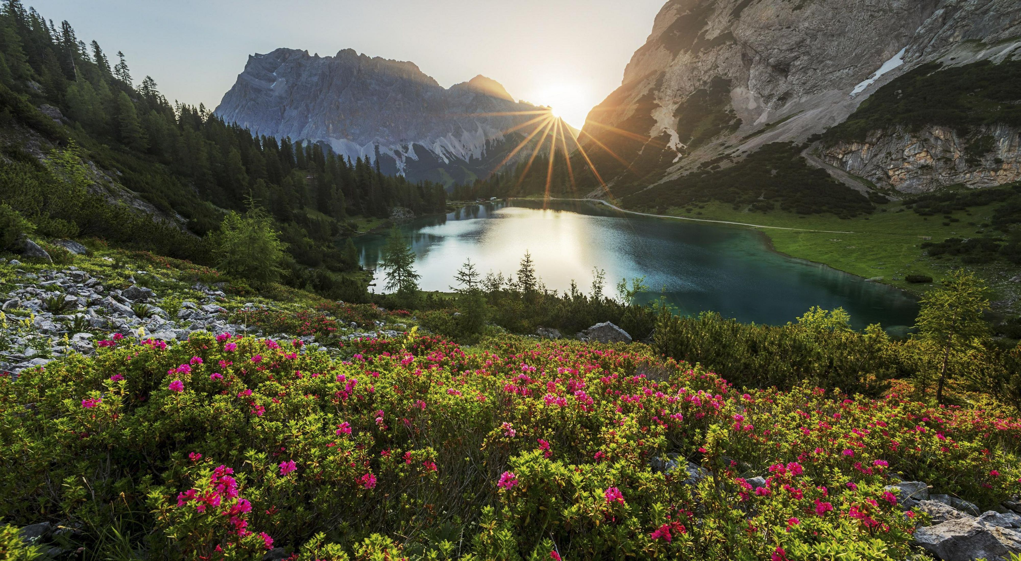 A wide, flat body of water is nestled in a mountain valley; the sun peeks through mountain peaks at the edge of the water.